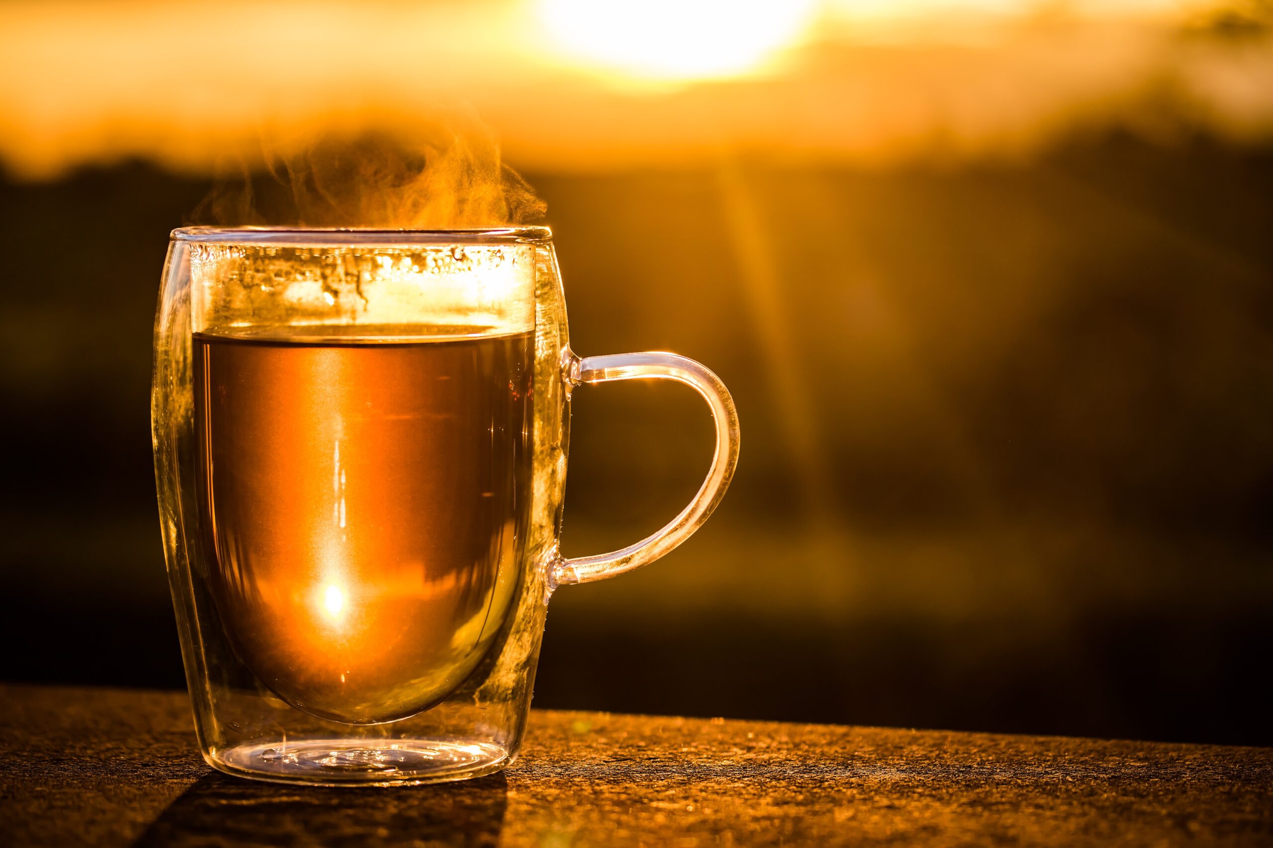 Doppelwandiges Glas mit dampfendem Tee seht im goldenen Abendlicht auf hölzernen Tischplatte.