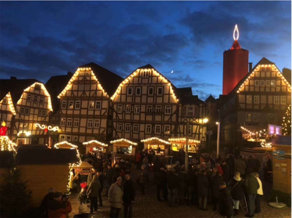 Schlitz Weihnachtsmarkt am Marktplatz bei Abenddämmerung: festlich beleuchtete Fachwerkhäuser, Buden und Besucher; im Hintergrund der riesige rote Kerzenturm.