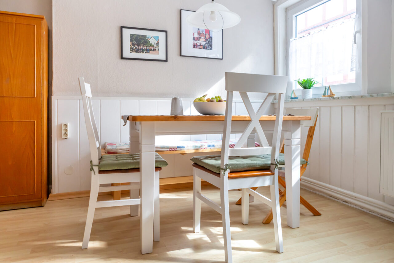 Esszimmer der Ferienwohnung mit weißem Holztisch, Stühlen, Obstschale und Wandbildern.