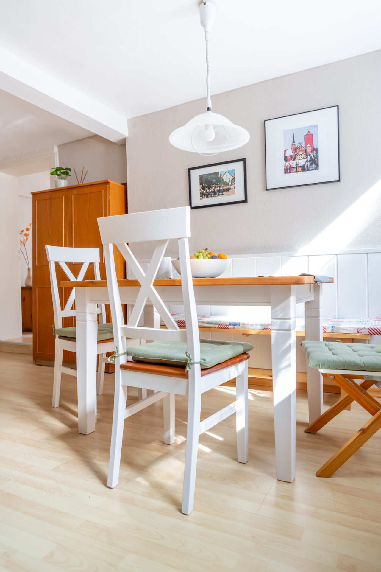 Esszimmer der Ferienwohnung mit weißem Holztisch, Stühlen, Obstschale und Wandbildern.